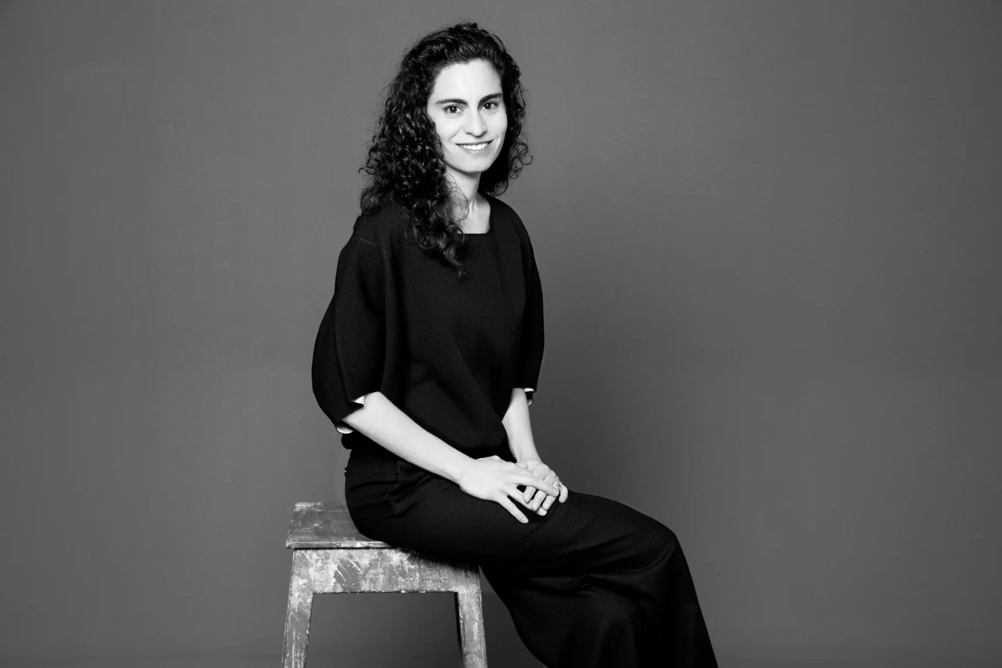 Black and white portrait of Yasmin Sabet sitting on a stool against a plain background
