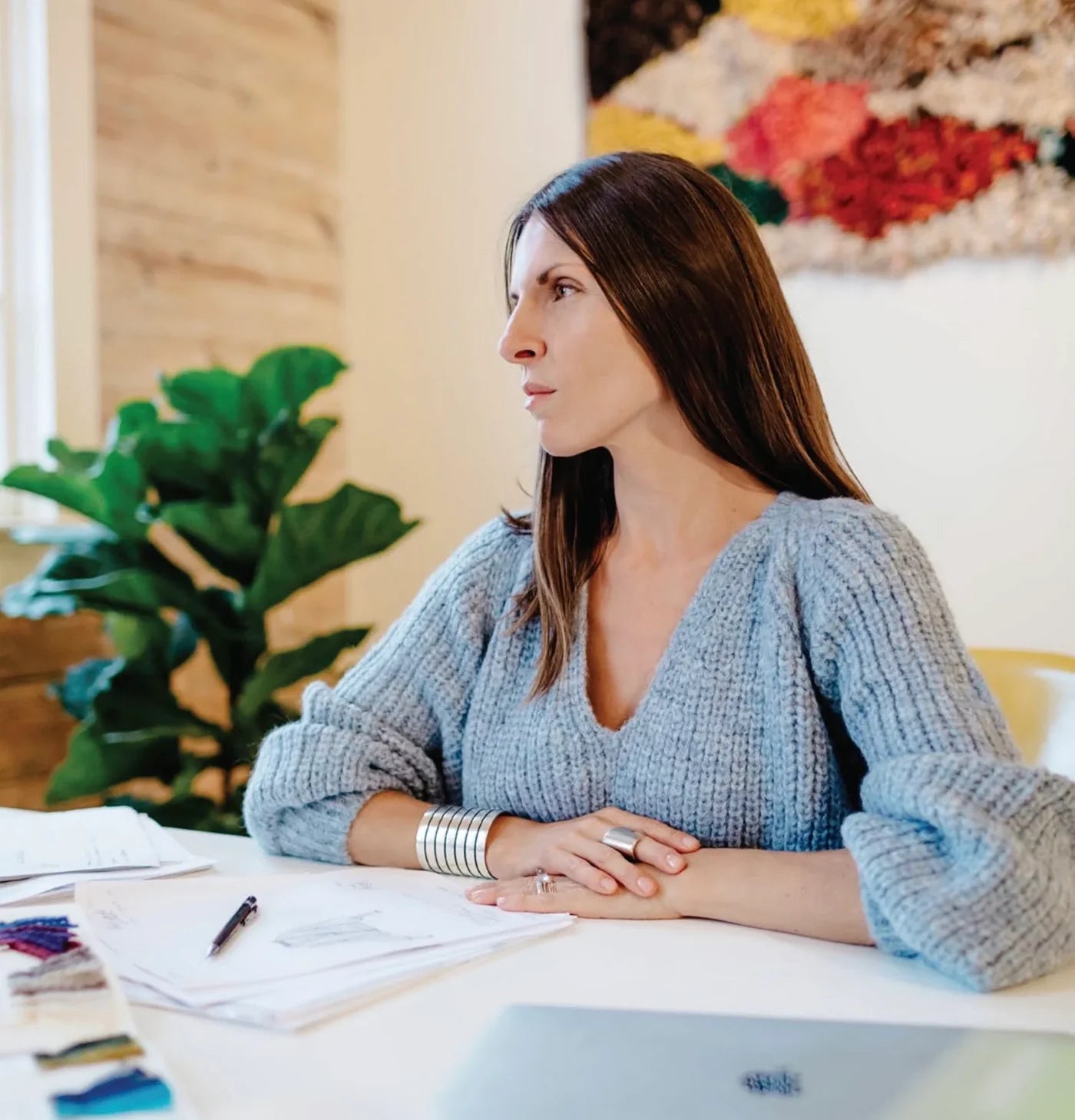 Woman sitting at a desk with papers and a laptop, wearing a blue sweater.